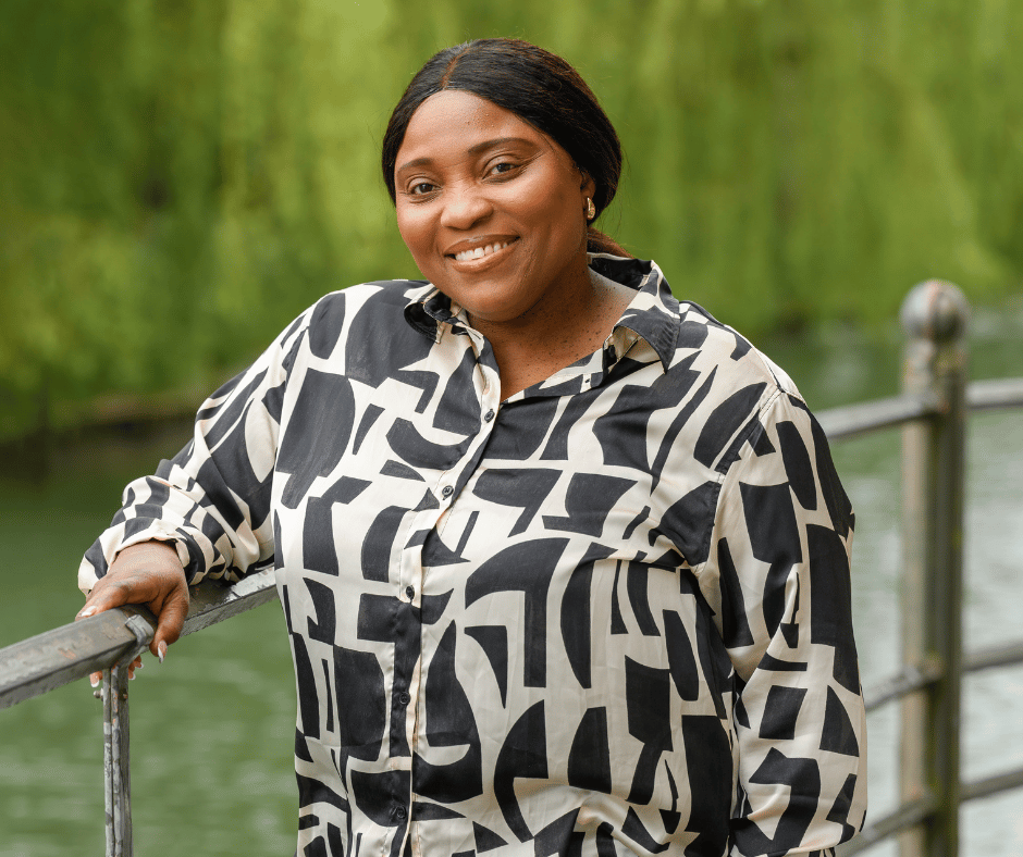 Liz Takyi - a black female with a white and black blouse standing against a railing smiling to the camera